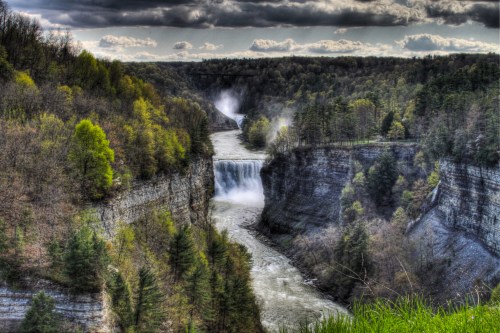 A scenic view of a waterfall cascading into a river, surrounded by lush green trees and dramatic clouds.