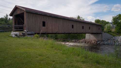 A wooden covered bridge spans a small river, surrounded by green grass and trees under a partly cloudy sky.