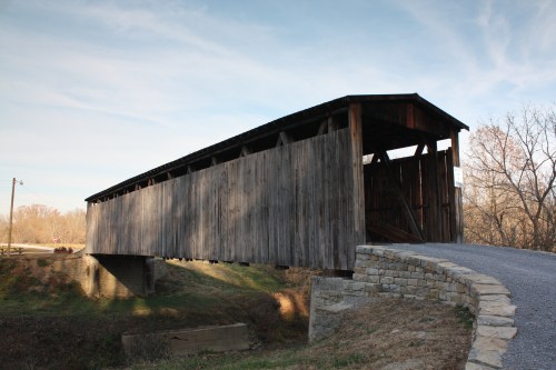 A wooden covered bridge spans a small creek, surrounded by trees and a gravel path under a clear blue sky.