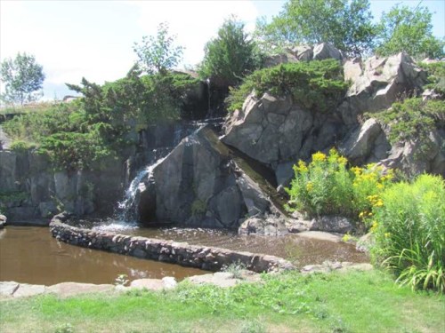 A serene landscape featuring a small waterfall cascading over rocks into a pond, surrounded by greenery and flowers.