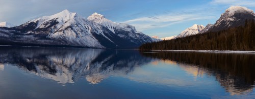 A serene lake reflects snow-capped mountains and a clear blue sky, surrounded by evergreen trees.