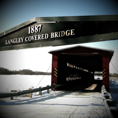 A historic covered bridge with a sign reading "Langley Covered Bridge 1887," surrounded by snow and a winter landscape.