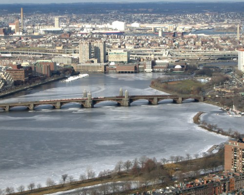 Aerial view of a frozen river with a bridge, surrounded by a cityscape and distant industrial areas.