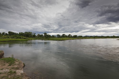 A calm river flows through a lush landscape under a cloudy sky, with greenery lining the banks.