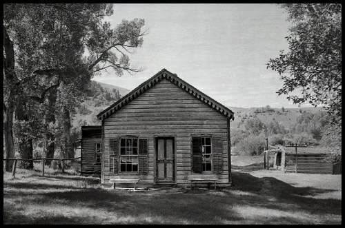 A black and white photo of a rustic wooden cabin surrounded by trees and hills.