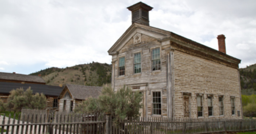 A weathered wooden building with a bell tower, surrounded by a fence and mountains in the background.