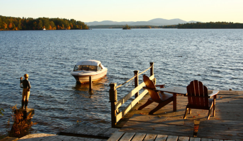 A serene lakeside scene at sunset, featuring a boat, a wooden dock, and a person fishing by the water.