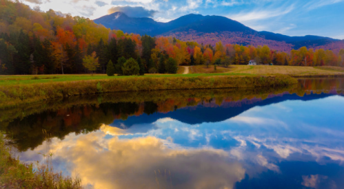 A serene landscape featuring colorful autumn trees, a calm lake reflecting mountains and a blue sky.