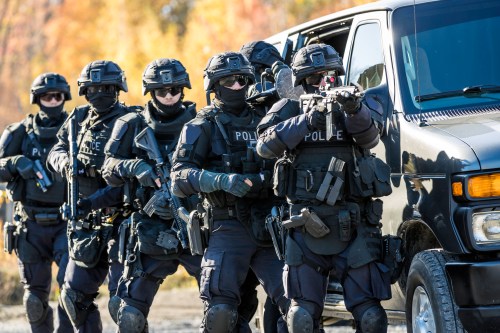 A group of heavily armed police officers in tactical gear approach a van during a training exercise.