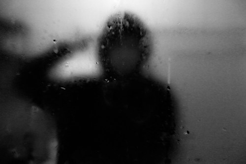 A blurred silhouette of a person with a hood, hand raised, behind a foggy glass surface.