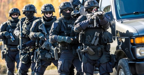 A group of heavily armed police officers in tactical gear approach a black van during a training exercise.