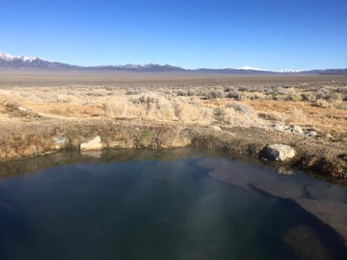A serene hot spring surrounded by dry grassland and distant mountains under a clear blue sky.
