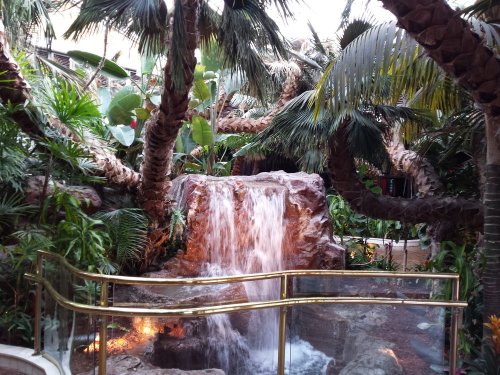 Lush indoor tropical scene featuring a waterfall surrounded by palm trees and vibrant greenery.