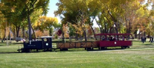 A small train with a blue engine and red passenger car travels through a park with colorful autumn trees.