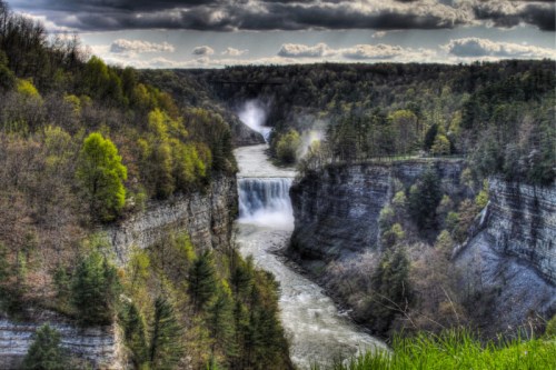 A scenic view of a waterfall cascading into a river, surrounded by lush green trees and dramatic clouds.