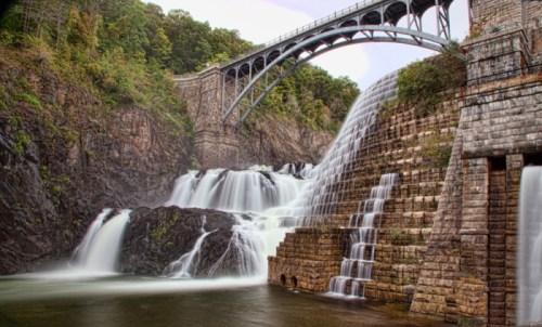 A scenic view of a waterfall cascading over stone structures, with a bridge arching above and lush greenery surrounding.