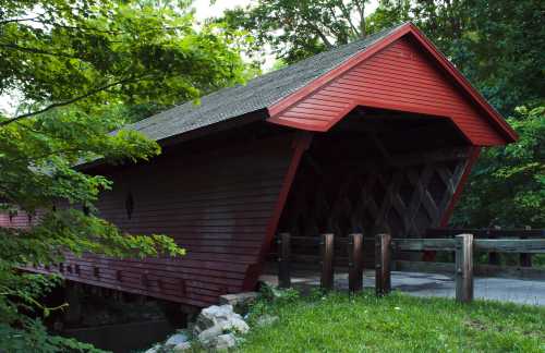 A red covered bridge surrounded by lush green trees, with a wooden railing in the foreground.