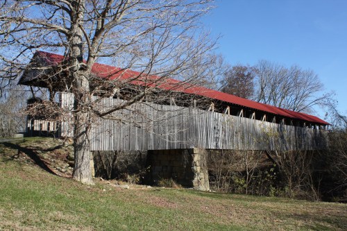 A rustic covered bridge with a red roof spans a small creek, surrounded by bare trees and a clear blue sky.