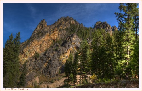 A rugged mountain peak rises above a forest of tall green trees under a clear blue sky.