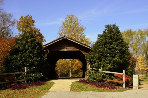 A wooden covered bridge surrounded by colorful autumn trees and shrubs, with a clear blue sky above.