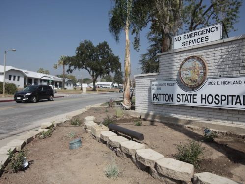 Sign for Patton State Hospital, with a clear sky and palm trees in the background, near a road. No emergency services noted.