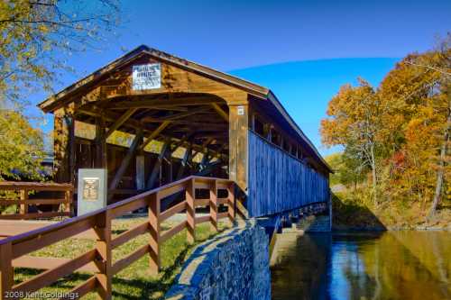 A wooden covered bridge spans a calm river, surrounded by autumn foliage and a clear blue sky.