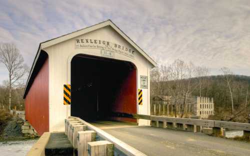 Rexleigh Bridge, a red covered bridge, spans a river with a cloudy sky in the background and an old building nearby.