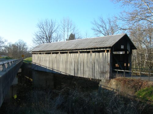 A wooden covered bridge spans a small creek, surrounded by bare trees and a clear blue sky.