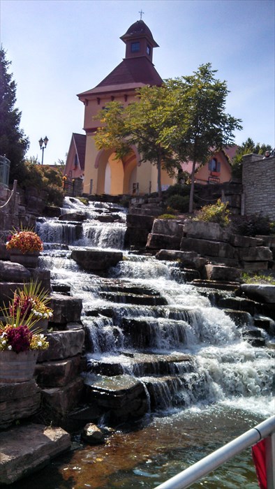 A picturesque waterfall cascades over rocks, with a charming building and trees in the background under a clear blue sky.