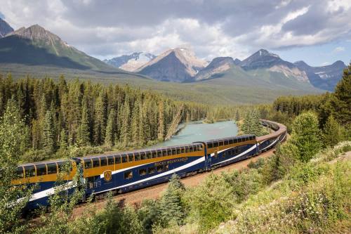 A scenic train winds through lush forests and mountains beside a river under a cloudy sky.