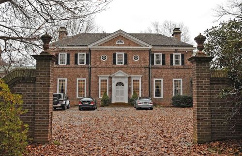 A large brick house with a symmetrical facade, surrounded by trees and a gravel driveway with parked cars.