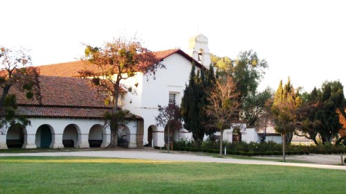 A historic building with a red-tiled roof, surrounded by trees and a grassy area, under a clear sky.
