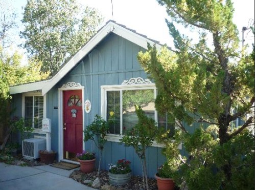 A charming blue house with a red door, surrounded by greenery and flower pots, on a sunny day.
