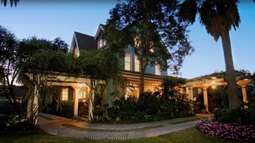 A charming two-story house surrounded by lush greenery and flowers, illuminated at dusk.