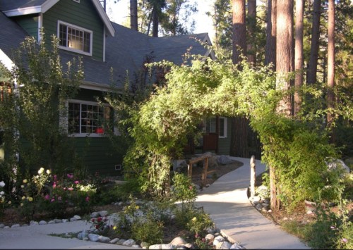 A charming green house surrounded by trees, with a flower garden and a stone pathway leading to an archway of greenery.