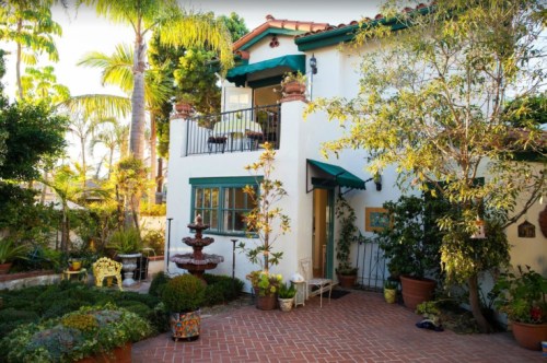 Charming white house with a balcony, surrounded by lush greenery and potted plants, featuring a small fountain in the garden.