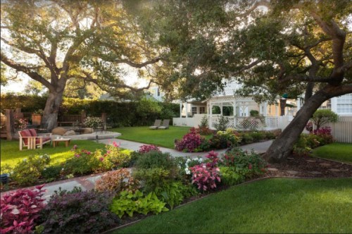 A serene garden with vibrant flowers, lush grass, and two large trees, featuring a cozy seating area and a white gazebo.