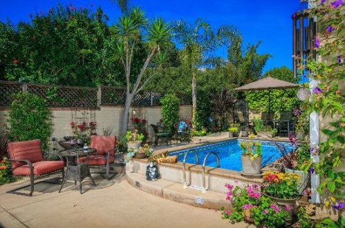 A vibrant backyard with a pool, surrounded by lush greenery, colorful flowers, and cozy seating areas.