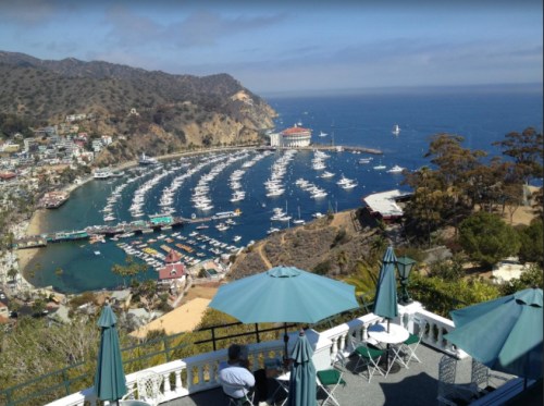 A scenic view of a coastal harbor with boats, surrounded by hills and a clear blue sky. Tables with umbrellas in the foreground.