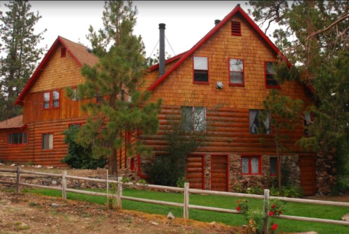 A large wooden cabin with a stone foundation, surrounded by trees and a grassy area, featuring red accents.
