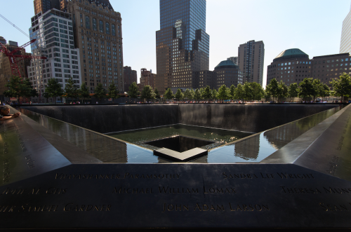A view of the 9/11 Memorial reflecting pool surrounded by trees and city buildings on a clear day.