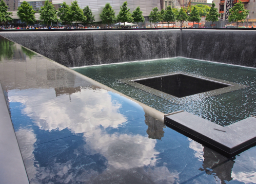 A reflective pool with a square void at its center, surrounded by trees and buildings, under a clear blue sky.