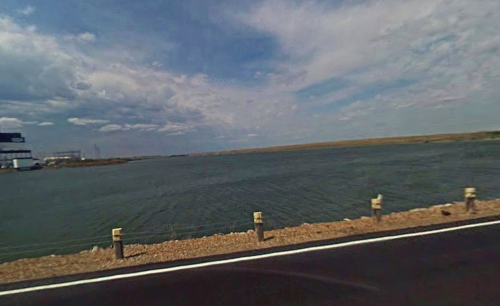 A scenic view of a calm body of water beside a road, with a cloudy sky and distant land in the background.