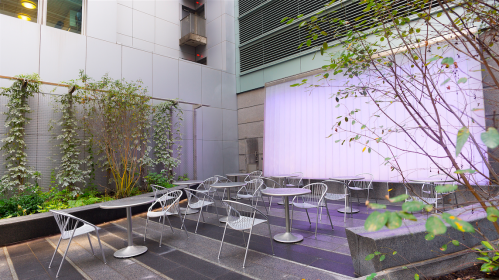 A modern outdoor seating area with metal chairs and tables, surrounded by greenery and a softly lit wall.