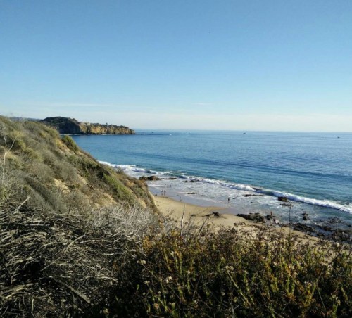 A serene coastal view with a sandy beach, gentle waves, and clear blue skies, framed by rocky cliffs and vegetation.