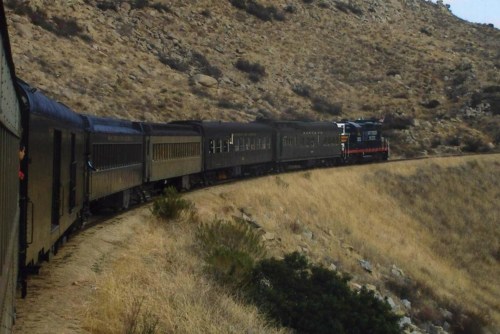 A train curves around a hillside, surrounded by dry grass and rocky terrain under a clear sky.