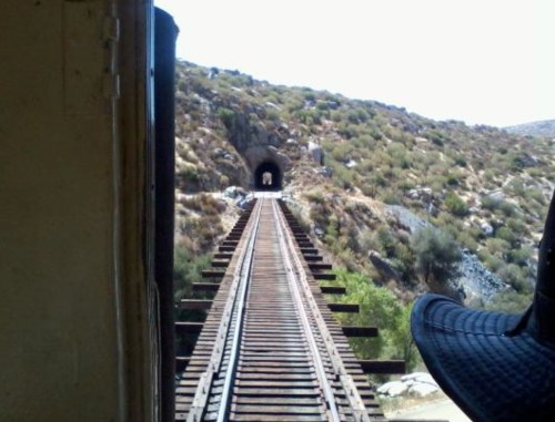 View from a train showing a railway track leading into a tunnel surrounded by hills and greenery.