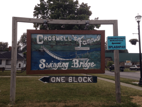 Sign for Croswell's Famous Swinging Bridge, indicating it's one block away, with additional info about a splash pad.