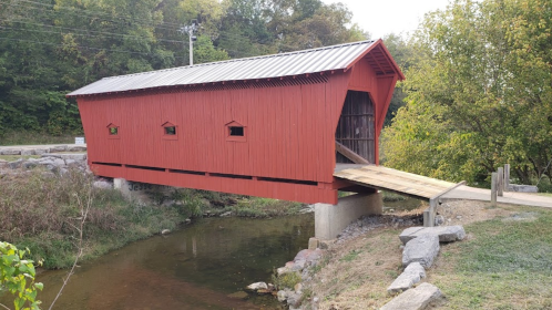 A red covered bridge spans a small creek, surrounded by greenery and a gravel path.