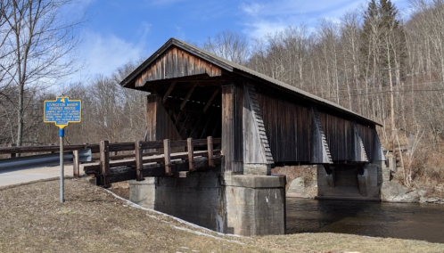 A wooden covered bridge spans a river, with a historical marker nearby and trees in the background.